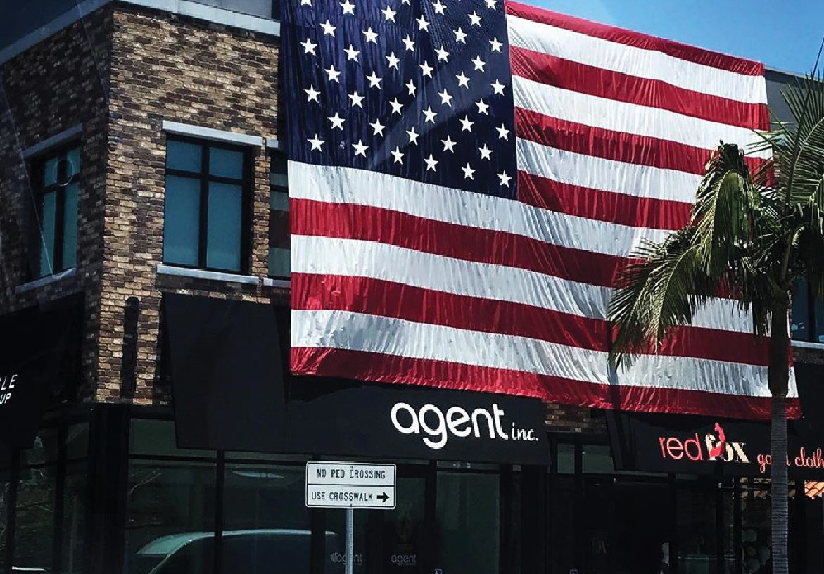 AGENT INC. logo displayed on the exterior of a modern real estate building, prominently featuring a large American flag.