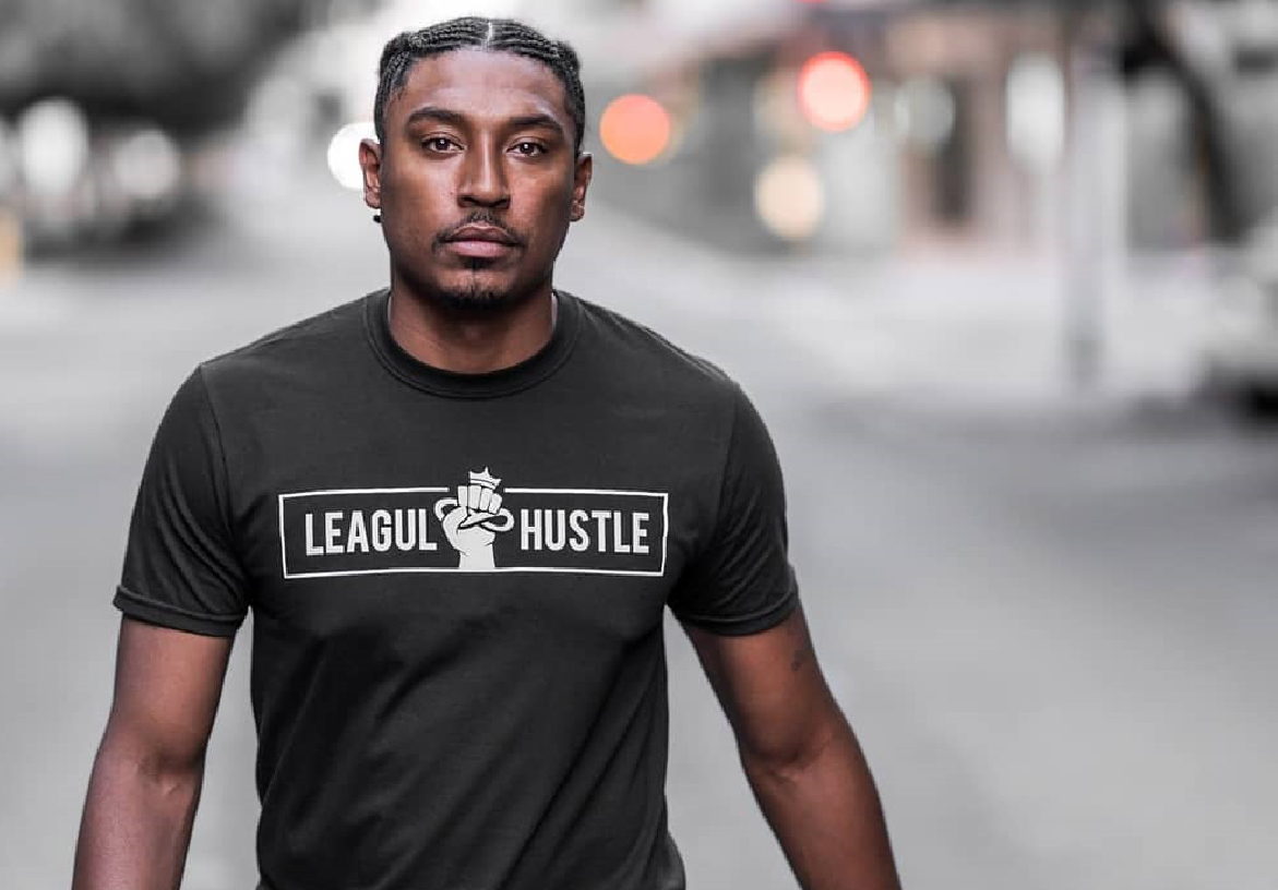 Male model walking on a city street wearing a black Leagul Hustle t-shirt with the white box logo, representing the brand's urban lifestyle aesthetic.
