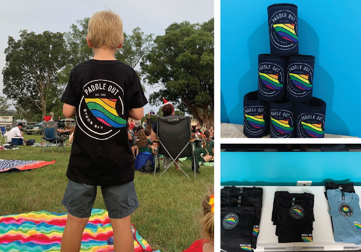 Paddle Out branded merchandise including black t-shirts and stubby holders displayed at an outdoor market event.