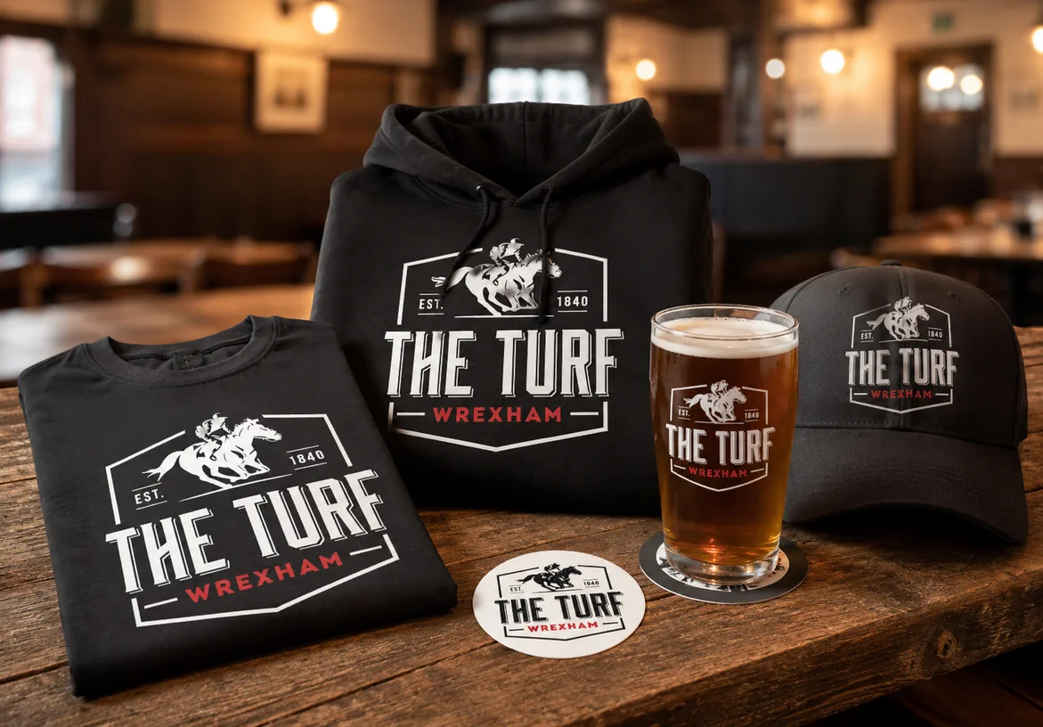 The Turf hoodie, pint glass, sticker, and merchandise arranged on a wooden pub table