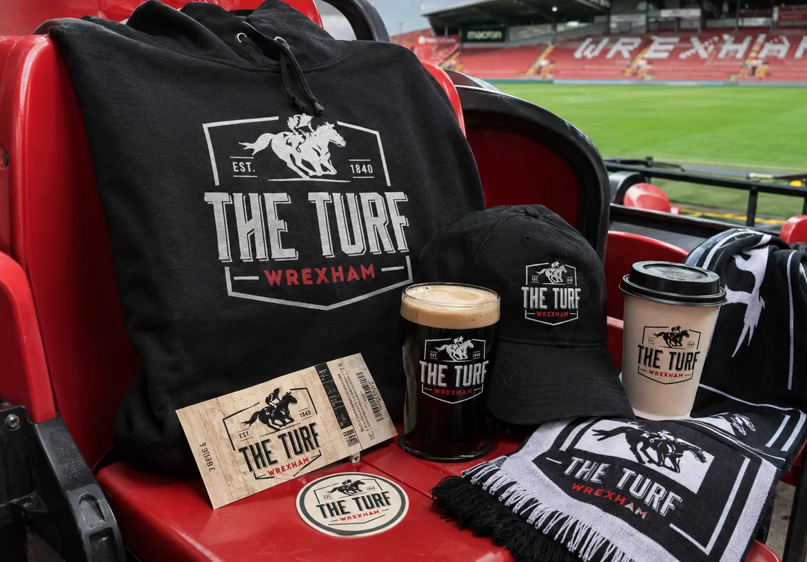 The Turf scarf, pint glass, coffee cup, and programme displayed on a stadium seat